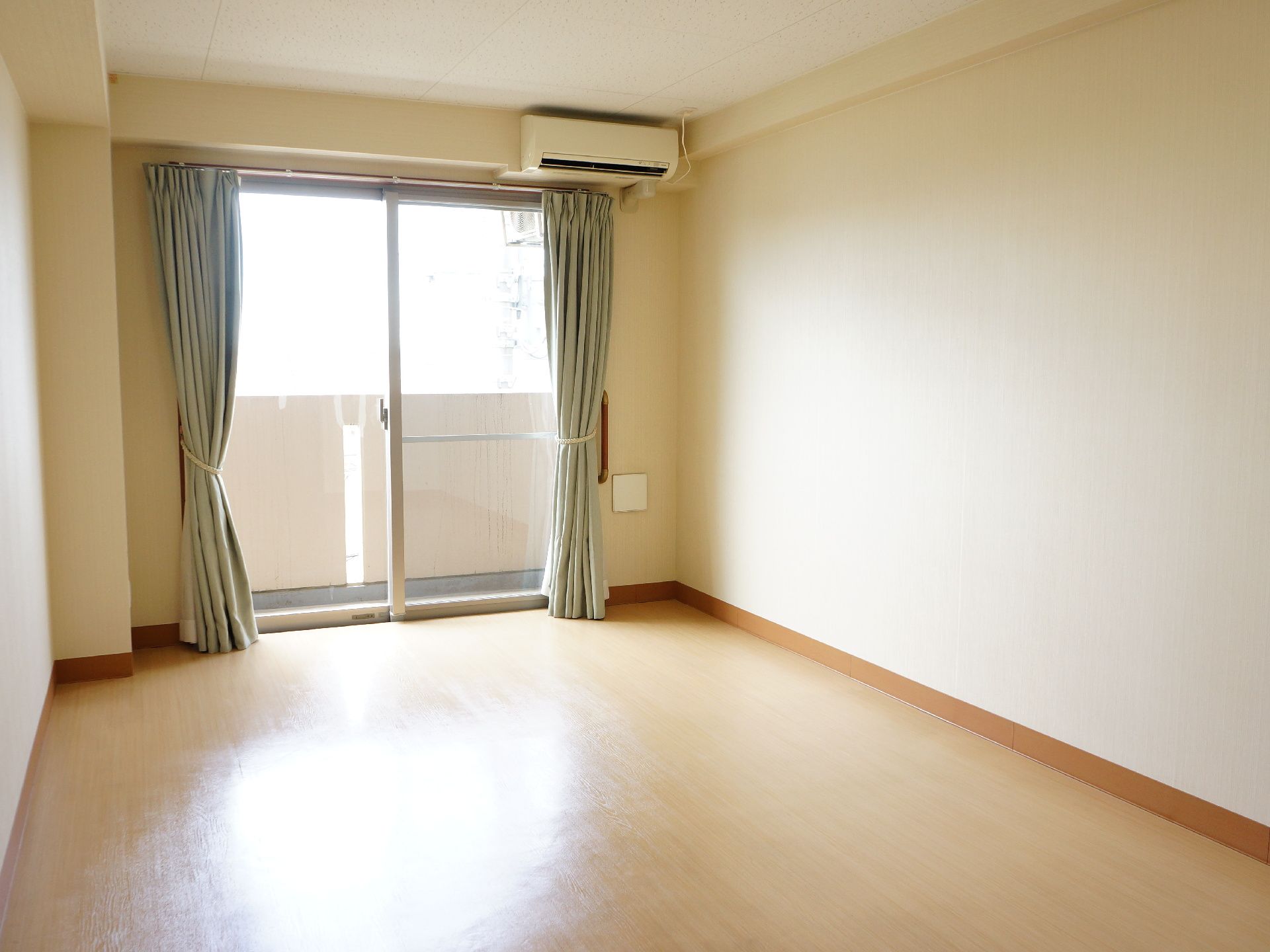 個室内には家具が一つも置いておらず空室状態で、上部にはエアコンが設置されており、窓からは光が差し込んでいる。