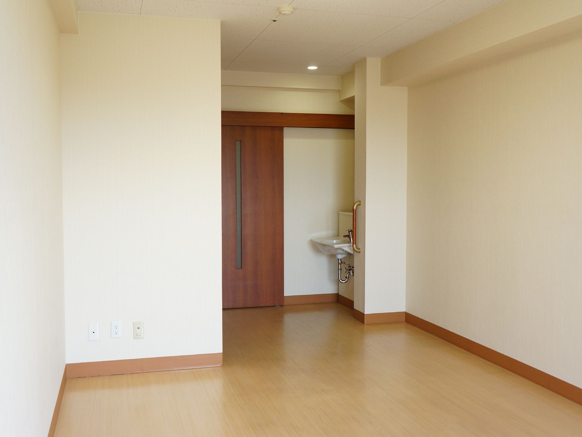 何もなく空室状態の個室の出口付近にはドアがあり、右隅には出っ張った壁に手すりと、隠れるように洗面台がある。