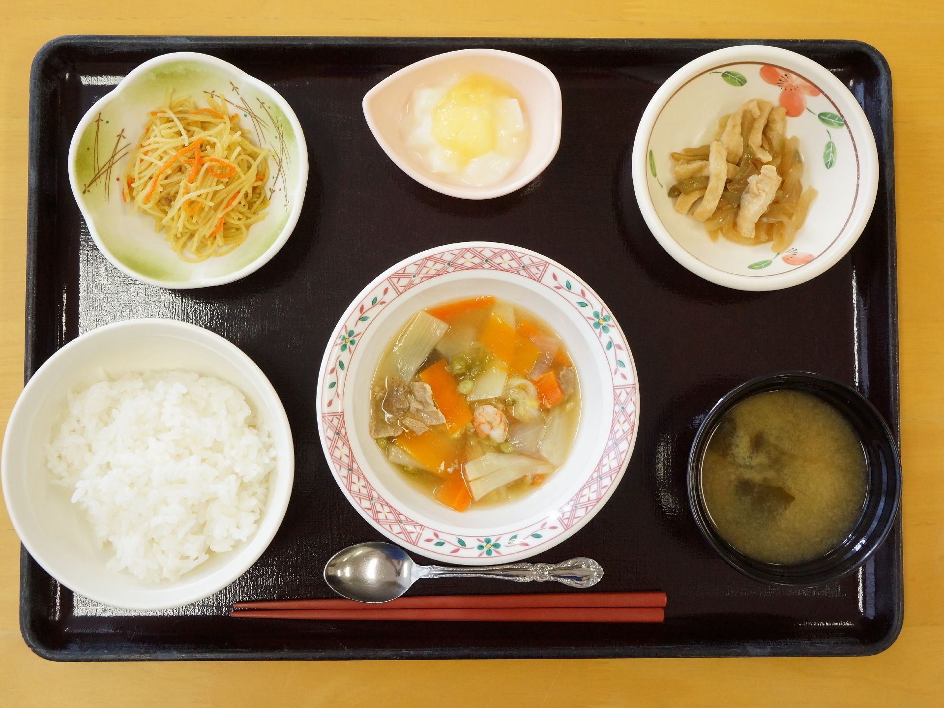 黒いお盆には6種類の食事が並んでいる。箸とスプーンが手前にあり、ご飯とみそ汁を中心に、おかずやデザートがそれぞれ小皿に入っている。