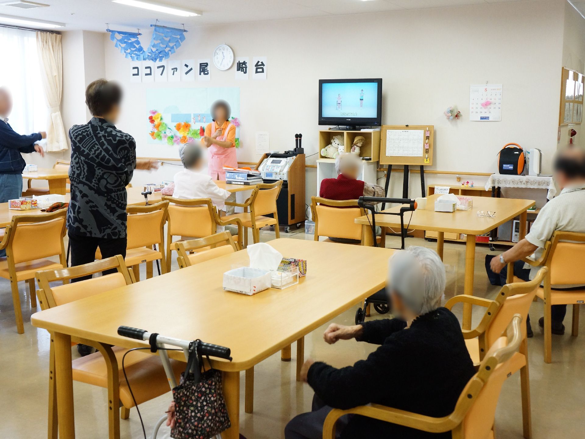 共有ルームでは、立った状態のスタッフ、立ったり座ったりしている入居者が、向かい合って運動をしている。