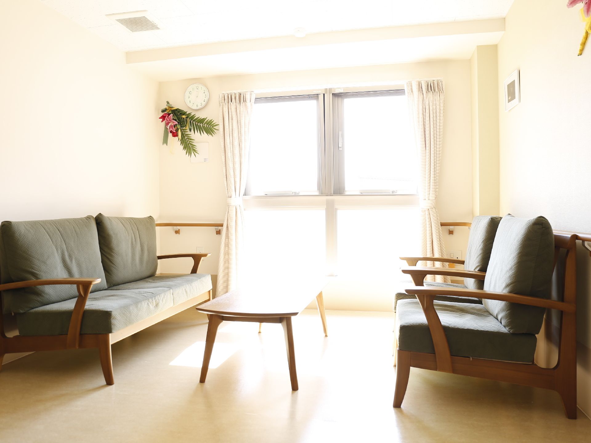 木目調のソファーと、木造りのテーブルがある。壁には時計が設置され、花も展示されている。窓からは明るい日差しが入り込んでいる。