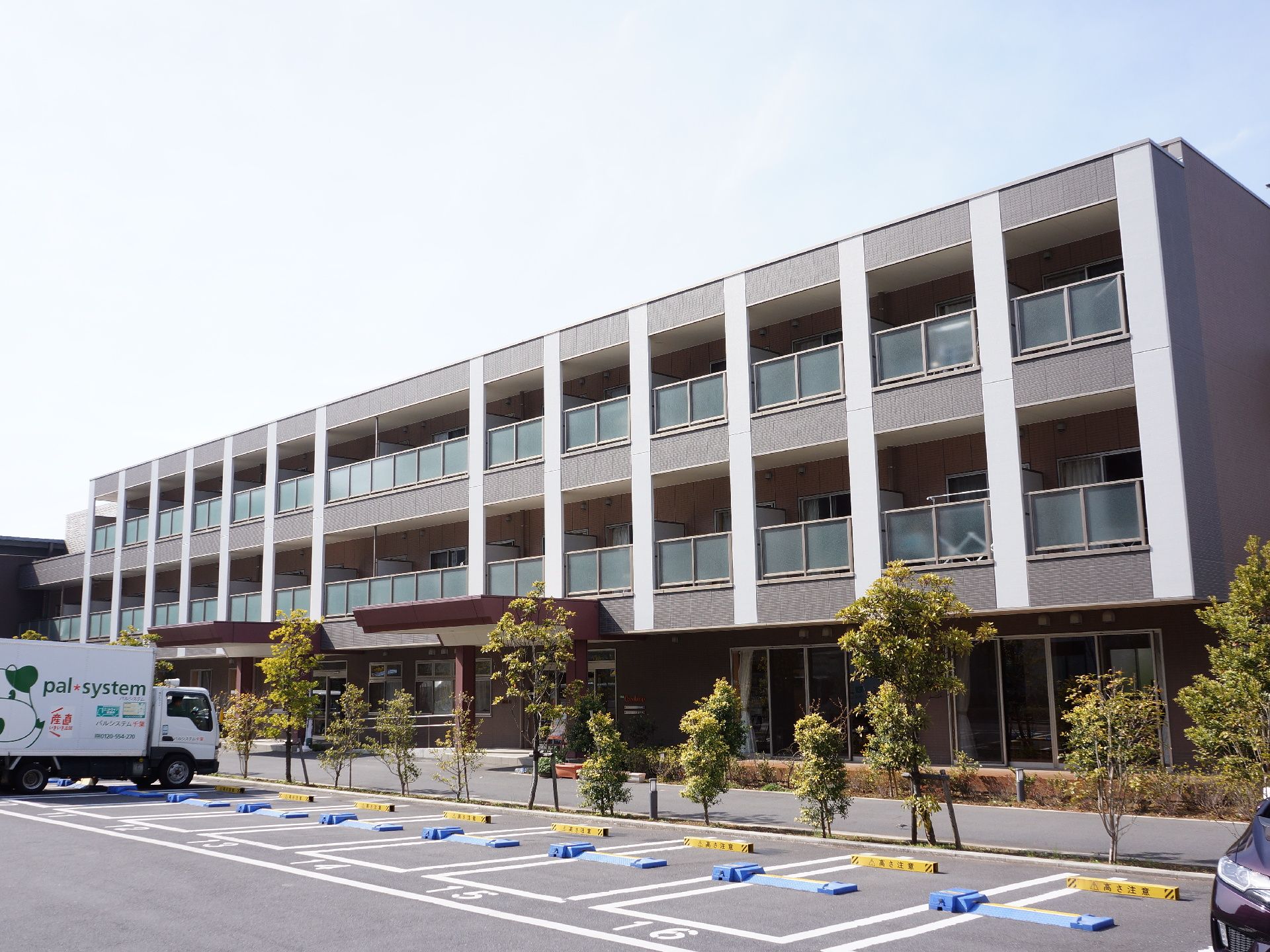 3階建ての茶色系統の建築物。正面玄関にはスロープがある。十数台の駐車スペースがある。駐車場はコインパーキング式になっている。
