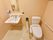 ベージュ色ベースの個室内に、手洗い場も一緒になっているタイプのトイレである。手洗い場は左側にあり、鏡や棚が付いている。