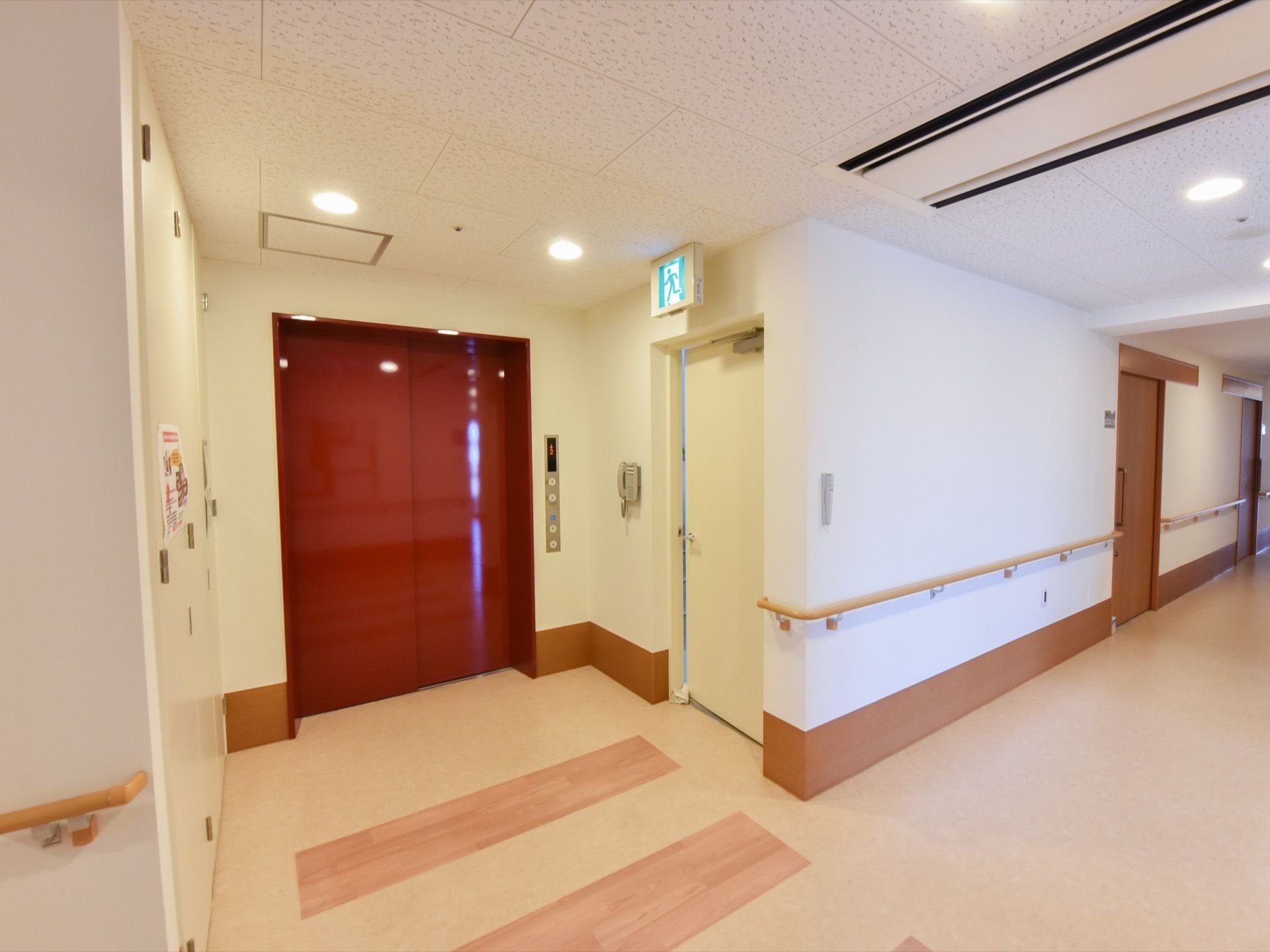 ワインレッド色のエレベーターの扉は、通路から奥まった場所にある。通路には、木製の手すりが取り付けられている。