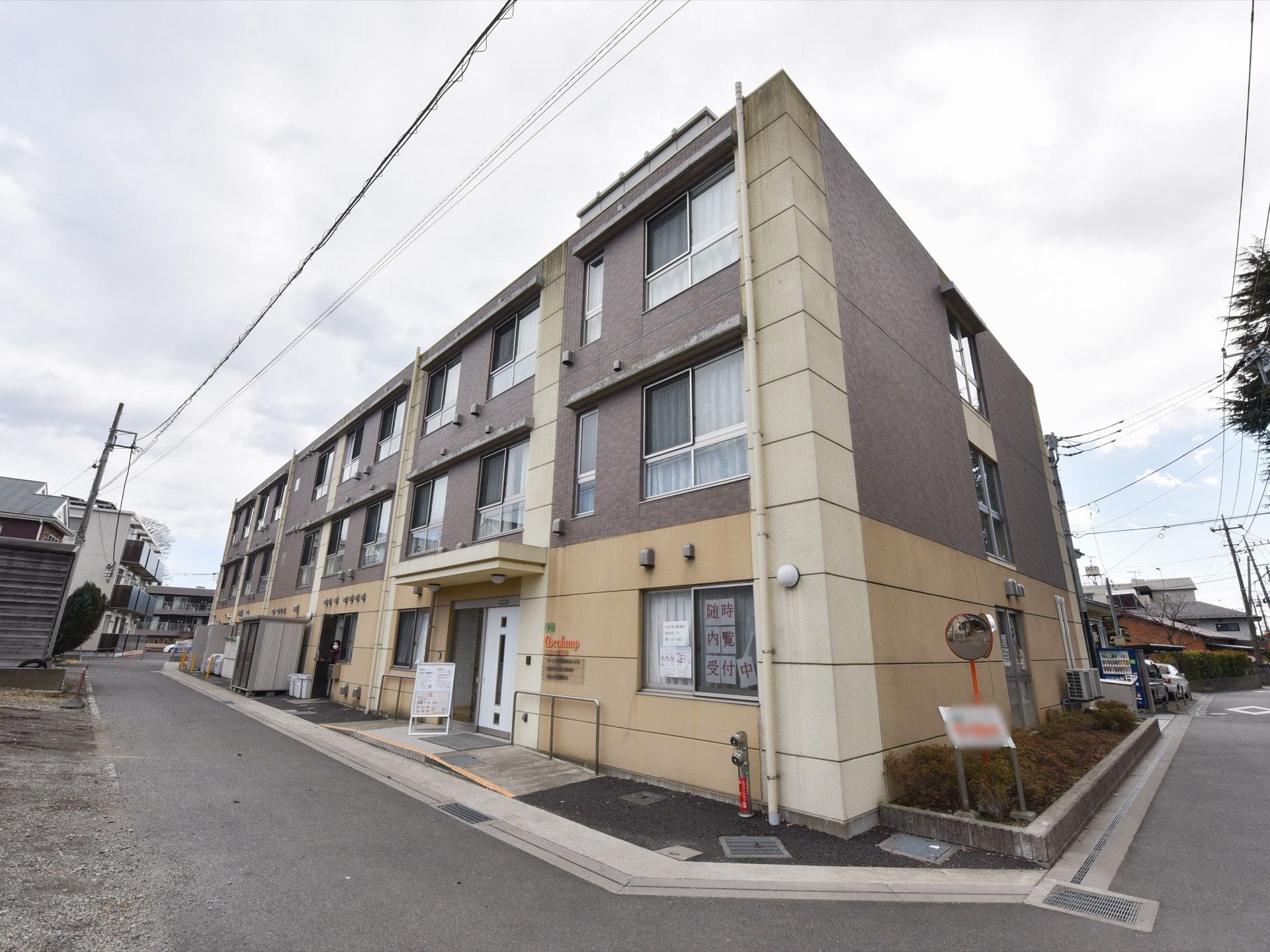 3階建ての施設の外観は、１階部分がベージュで２・３階部分はグレー系のツートンカラーである。窓には内覧受付中のチラシが貼られている。