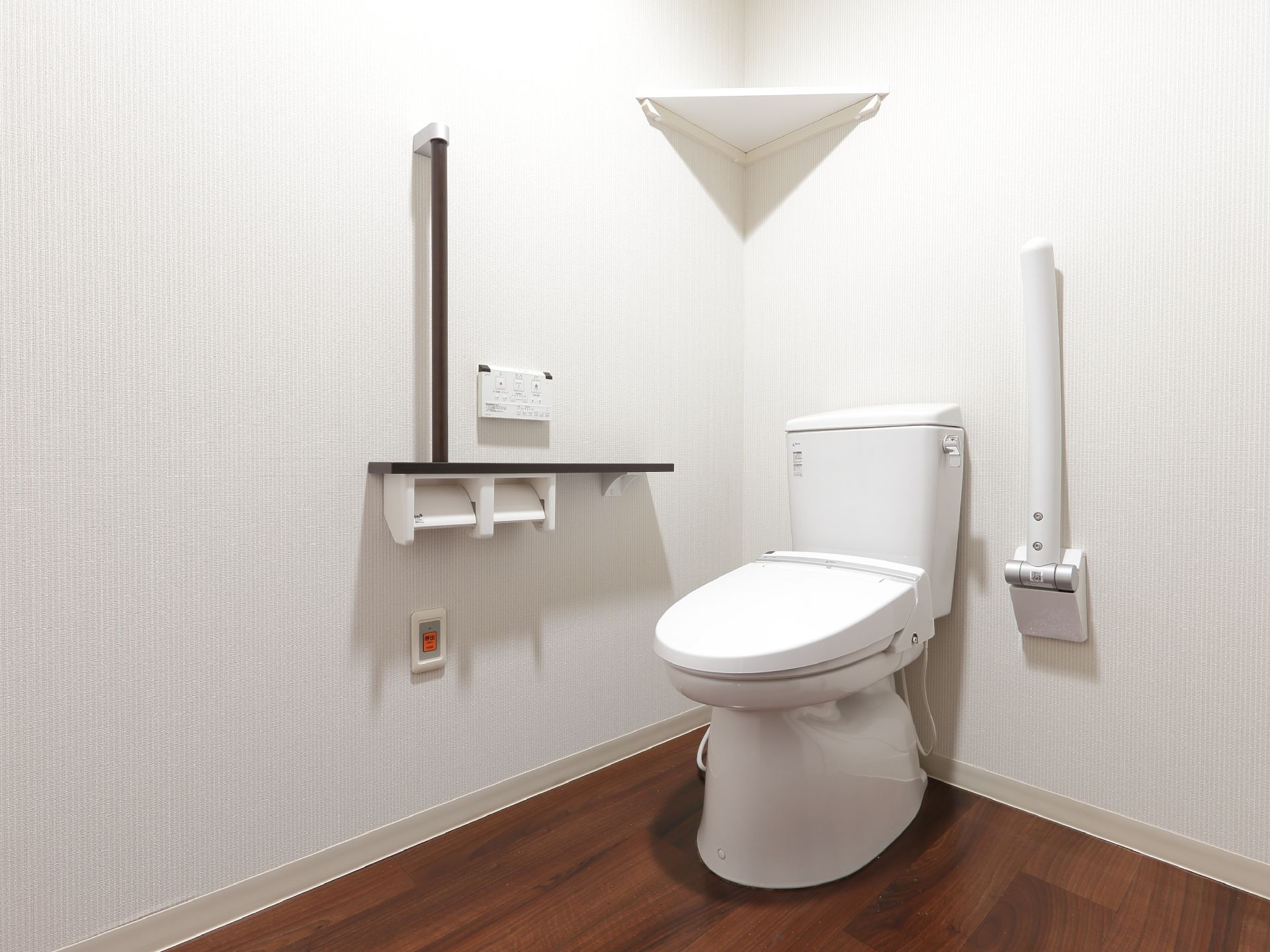 トイレは洋式で、清潔感がある白色。手すりと反対側に可動式の手すりが設置されている。壁は白、床は茶色。