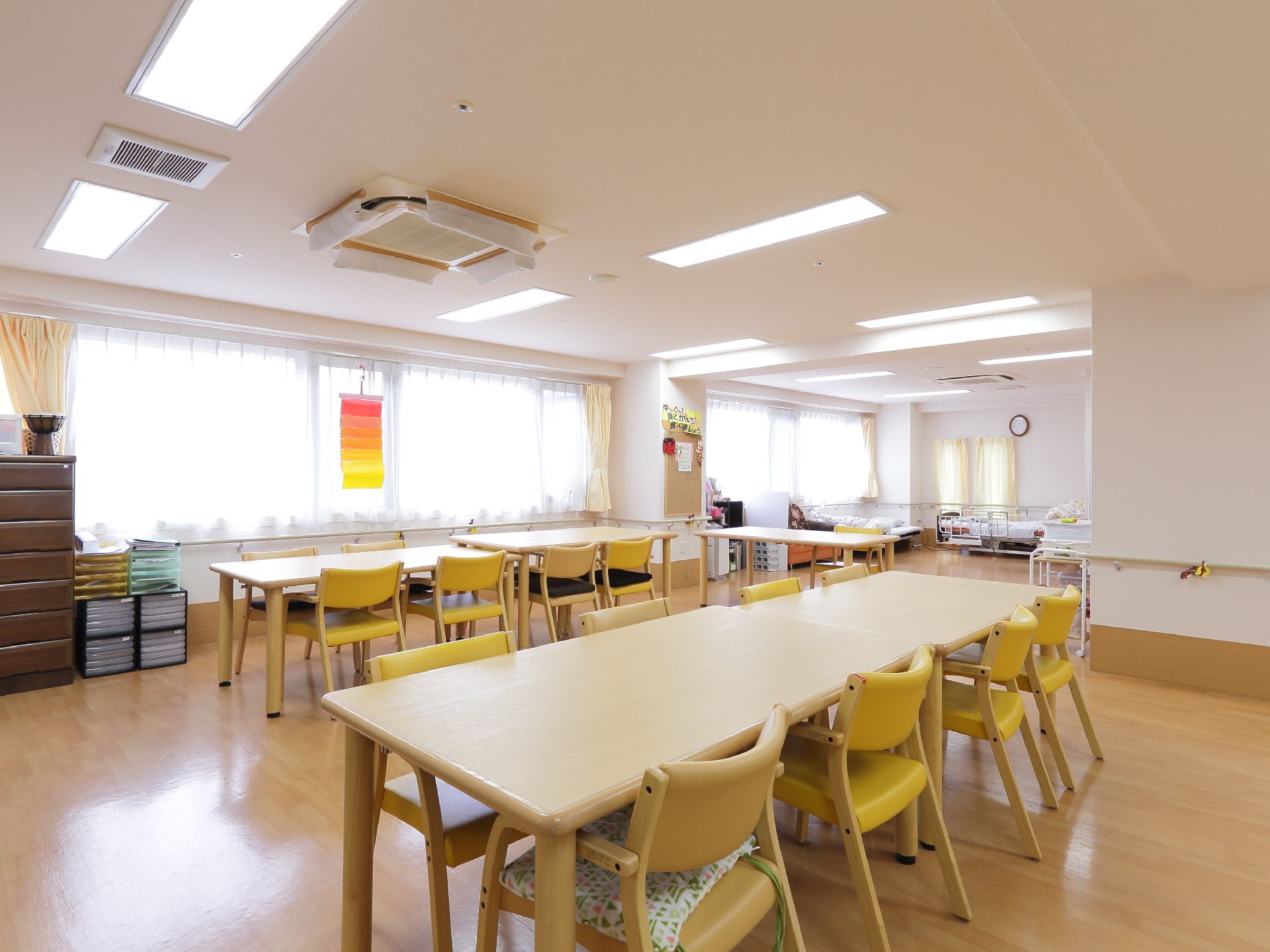 ナチュラル系の色のテーブルと椅子がたくさん置いてある。背もたれや座面が黄色なので部屋が明るく見える。