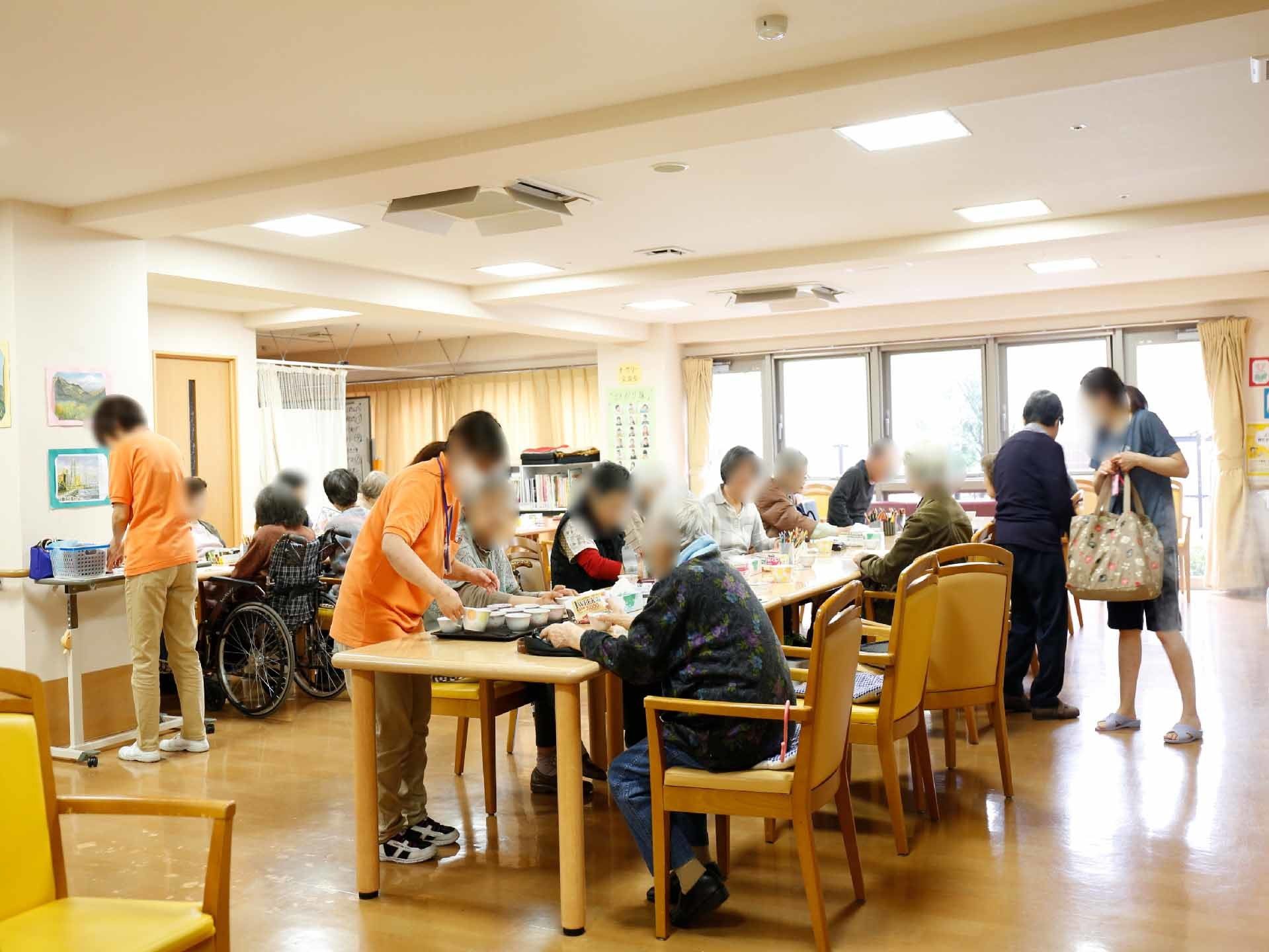 食事の様子。15名くらいの方が席についている。職員がお茶を入れて利用者さんが手伝おうとしているように見える。