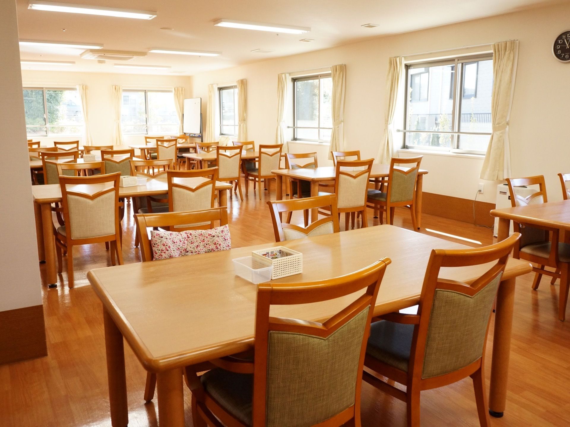 窓が多数設置されていて、明るい日差しが入っている。その空間にテーブルや椅子が並んでいる。椅子にクッションなども置いている。