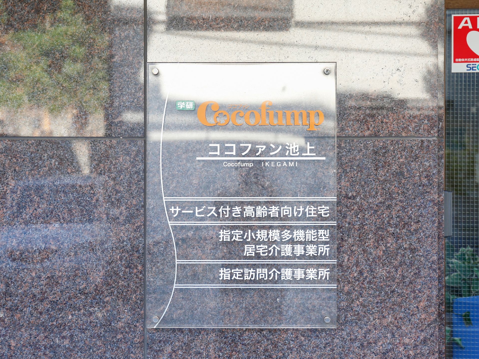 施設の入口には、透明のプレートが設置されている。オレンジ色の文字で施設名ロゴ、白い文字で施設内容が書かれている。