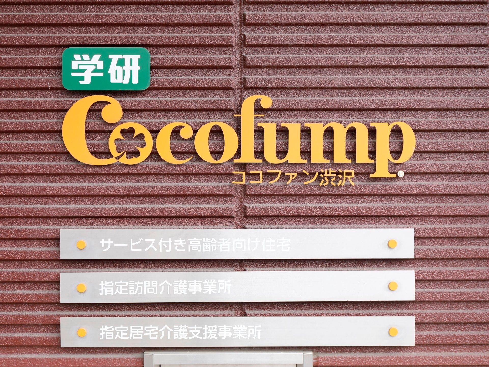 茶色い壁に緑と白とオレンジ色で書かれた施設の看板がある。施設名の下には施設についての説明が３行書かれている。