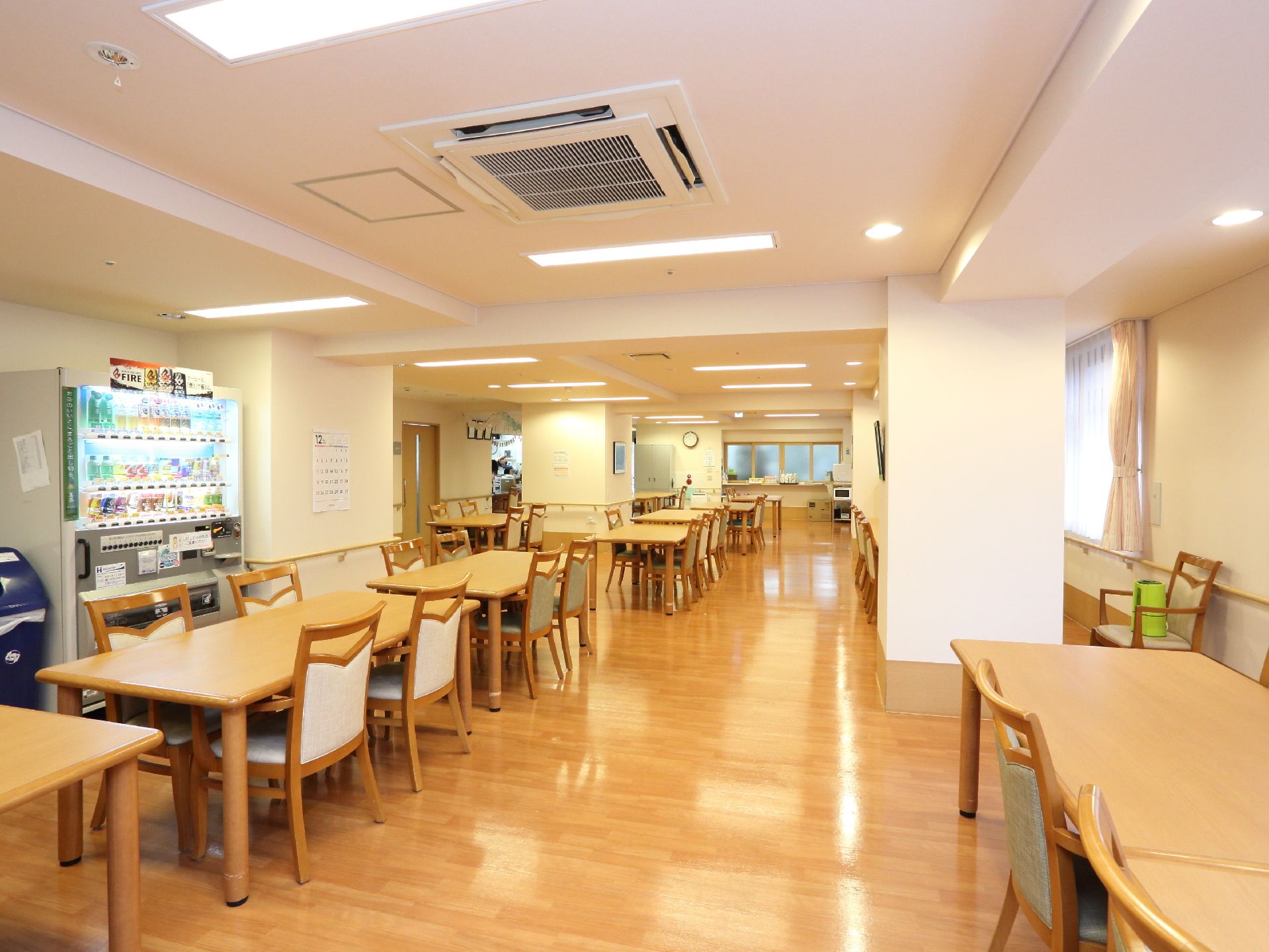 リビングの一角には自動販売機が設置され、隣にゴミ箱も置かれている。壁には手すりがあり、カレンダーなども貼られているのが見える。