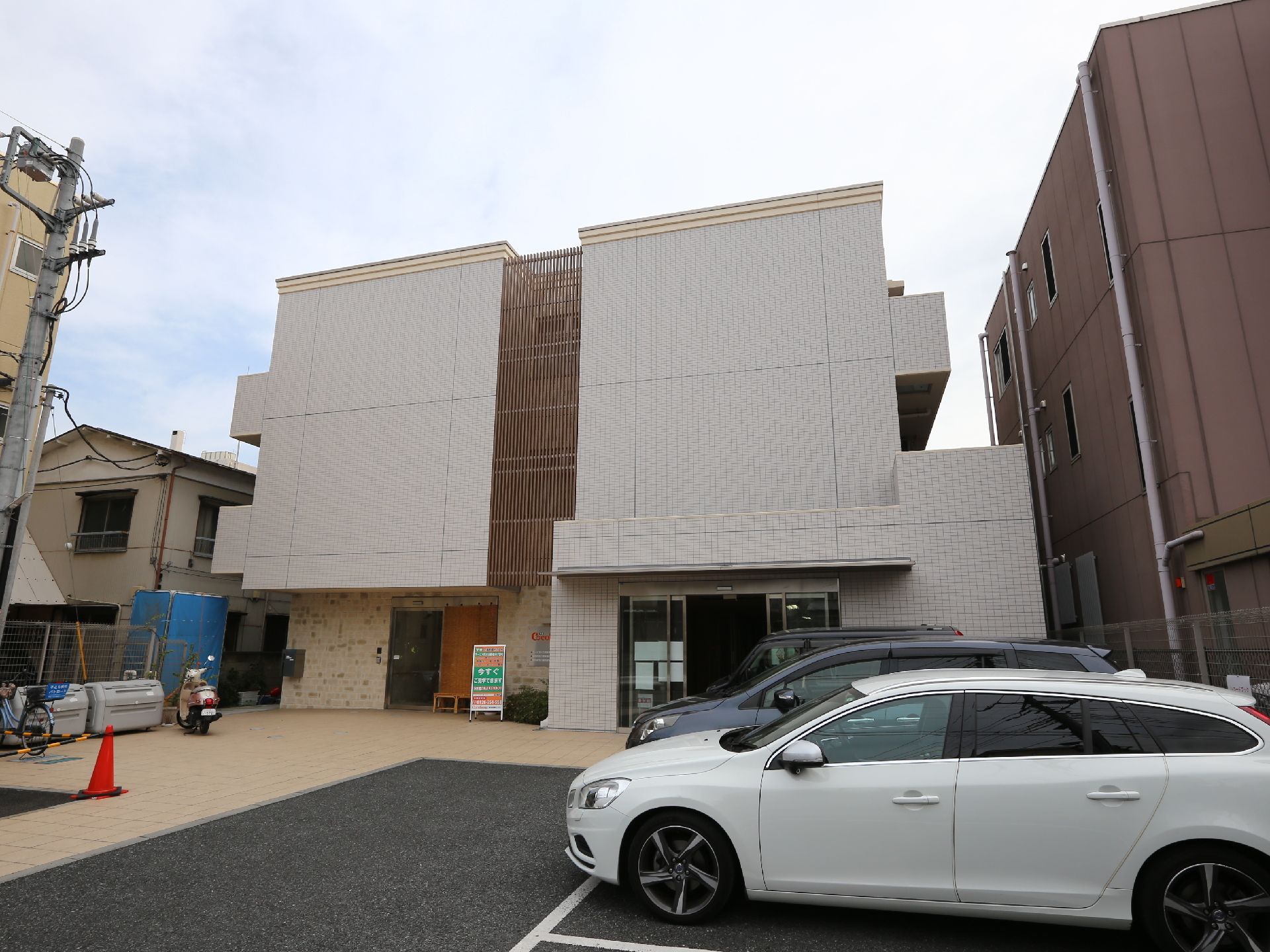 施設の外観はマンション風の佇まいの三階建の建物。敷地の手前は駐車場になっていて乗用車が複数台駐車しているのが写されている。