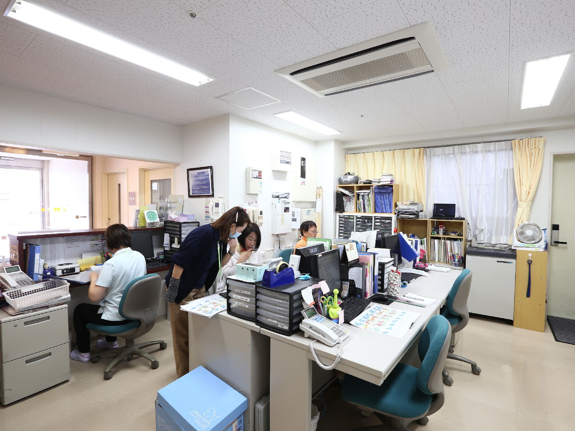 事務所はカウンターや受付も兼ねていて奥には出入り口のドアが見える。複数の職員が事務仕事や対応を行っている様子が写っている。