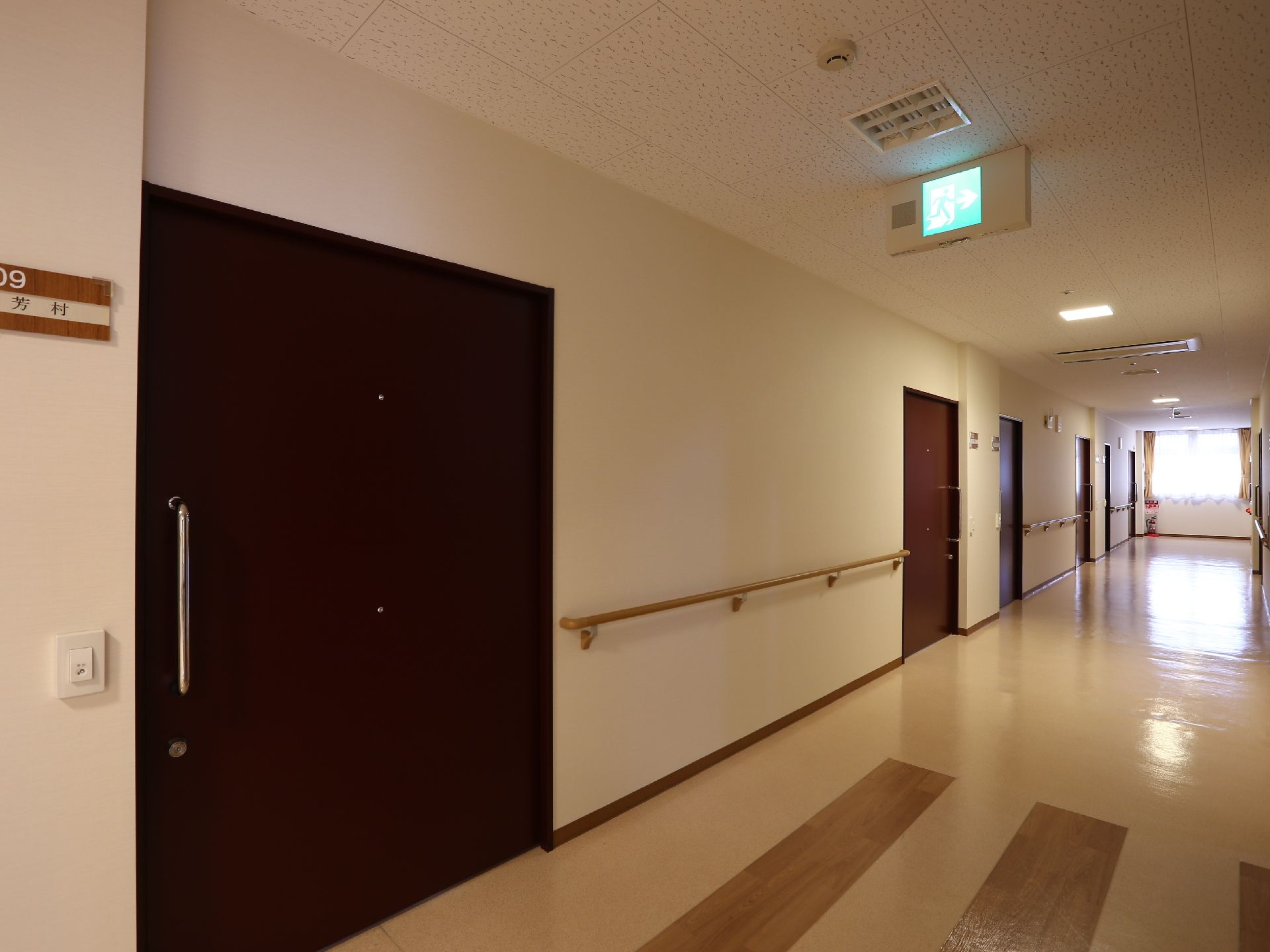 居室前の廊下も幅広に作られている。茶色の扉が奥まで並んでいて突き当たりは窓になっている。居室のドアの横には部屋番号のプレートがある。
