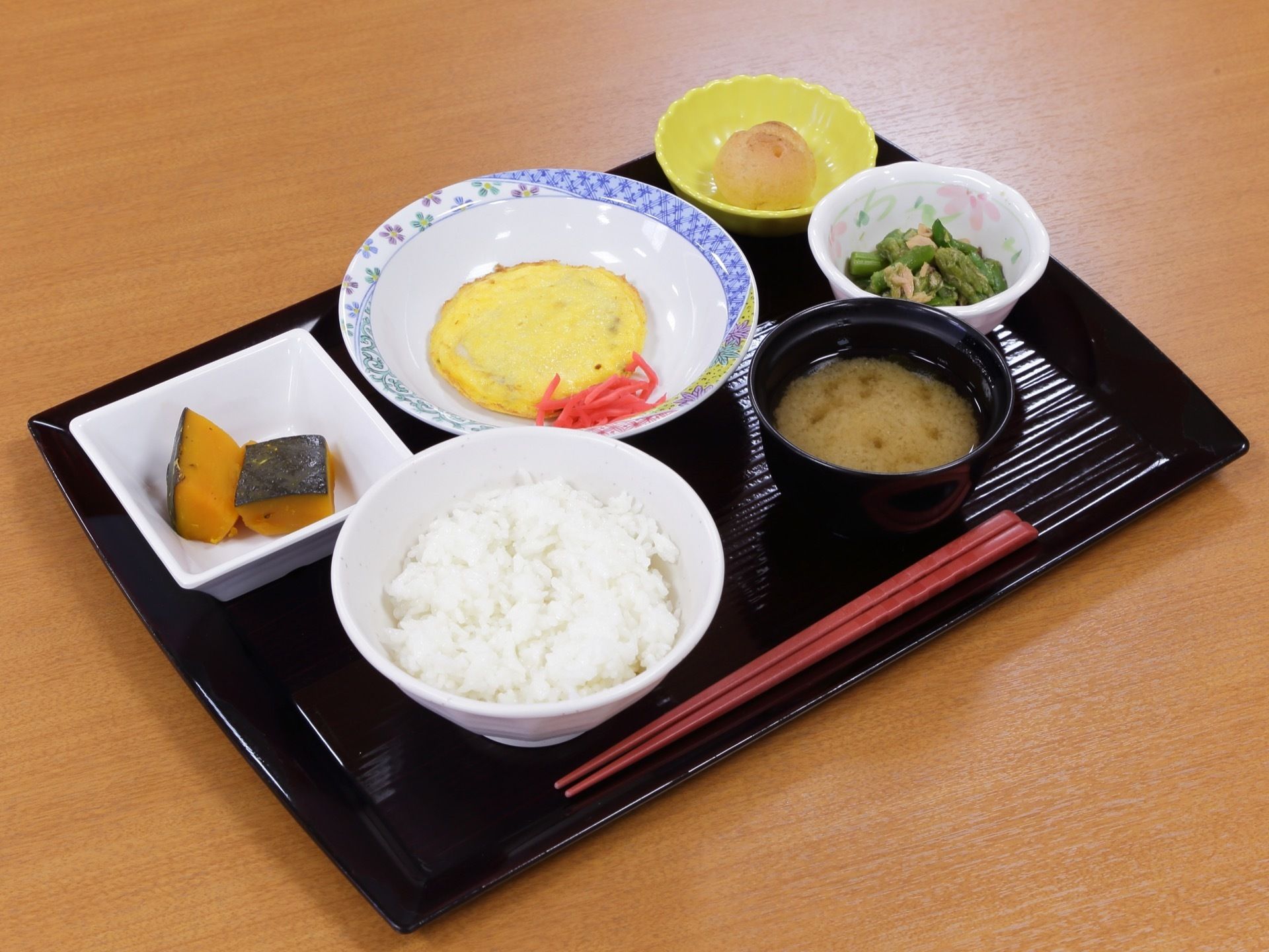 お盆に食事がセットされている。左に白いご飯、右に味噌汁、手前に箸がある。中央にメインの料理、副菜、小鉢、デザートもある。