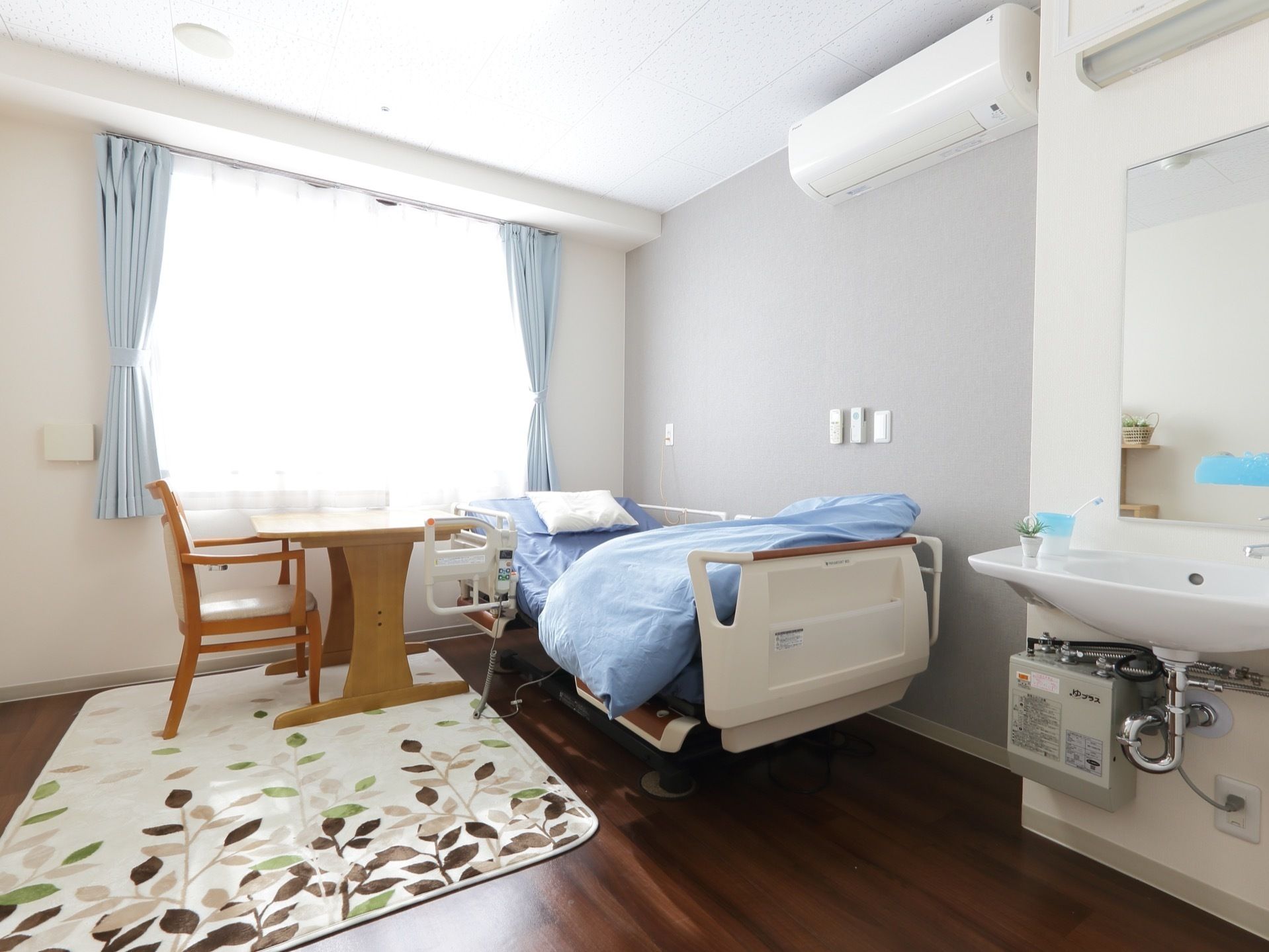 窓にはカーテンがあり、電動介護ベッドと家具、敷物、エアコン、洗面台が設置されている。壁にはリモコン、ナースコールがある。