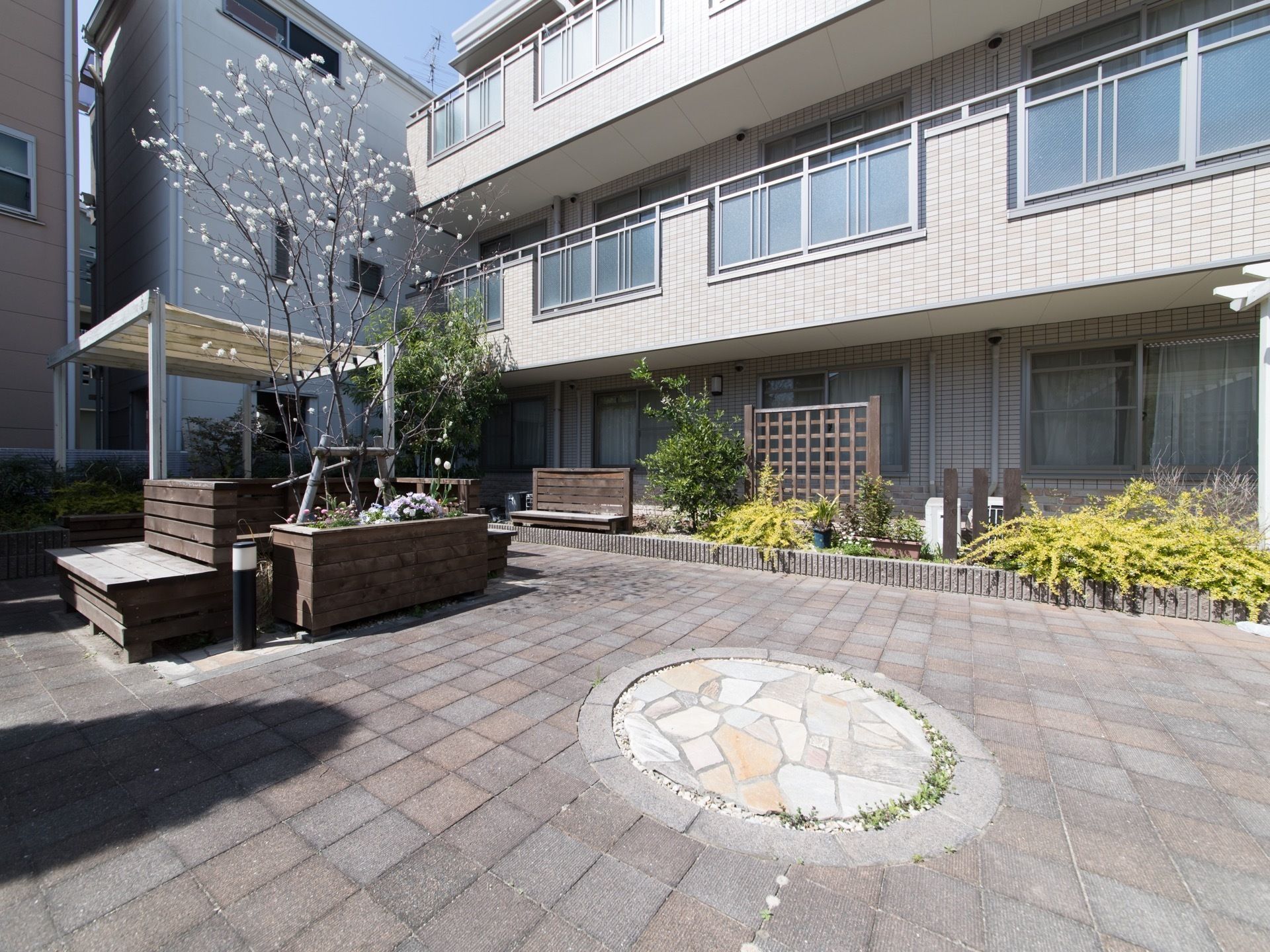 敷地内の建物横に設けられたテラスである。庭木が植えられた花壇があり、木製のベンチを設置している。自然豊かな空間である。
