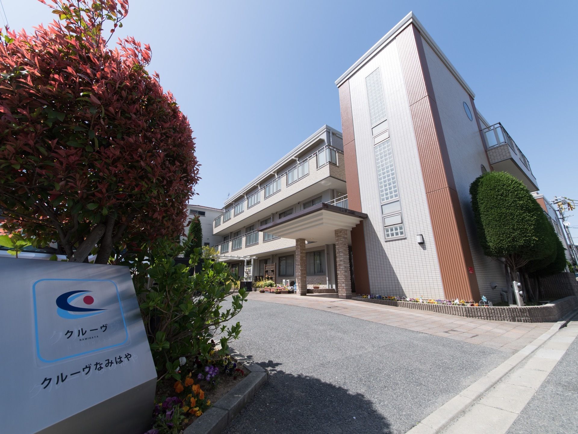 道路に面した敷地内の出入口の傍に、植栽の花壇を設け、その中に施設のロゴマークと施設名が書かれた看板を掲げている。