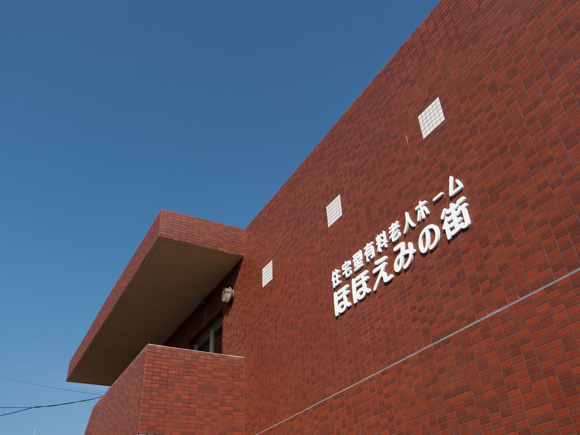 赤茶けたレンガ風の外壁の中に、白い文字で施設名のロゴが取りつけられている。写真奥には屋外階段がある。