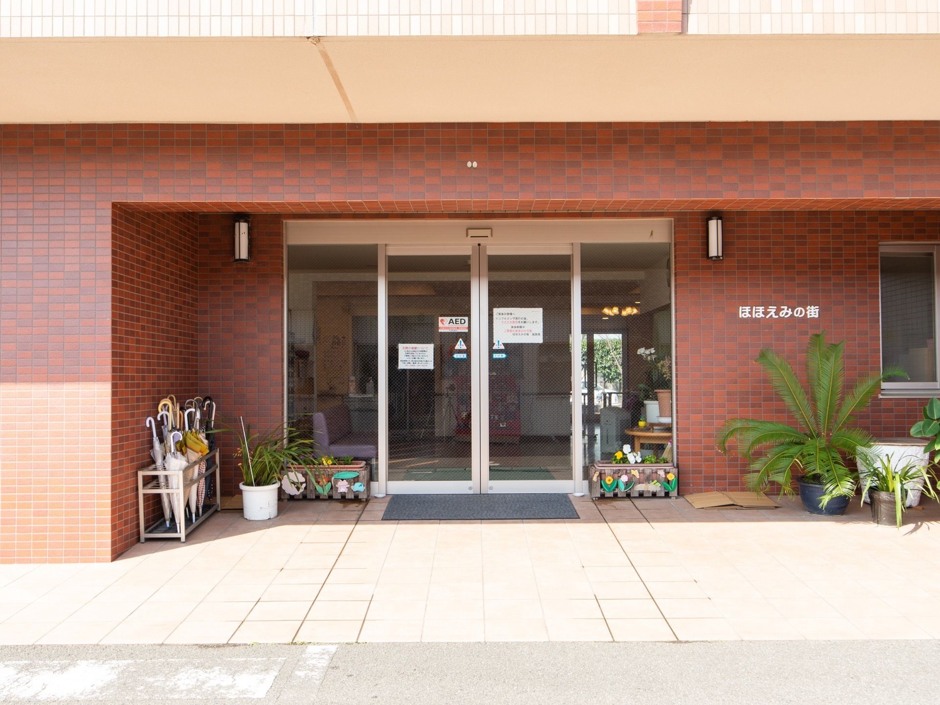 施設の正面玄関。赤レンガ風の壁の中にガラス張りの自動ドアが設置されている。周りには傘立てや観葉植物などが置かれている。