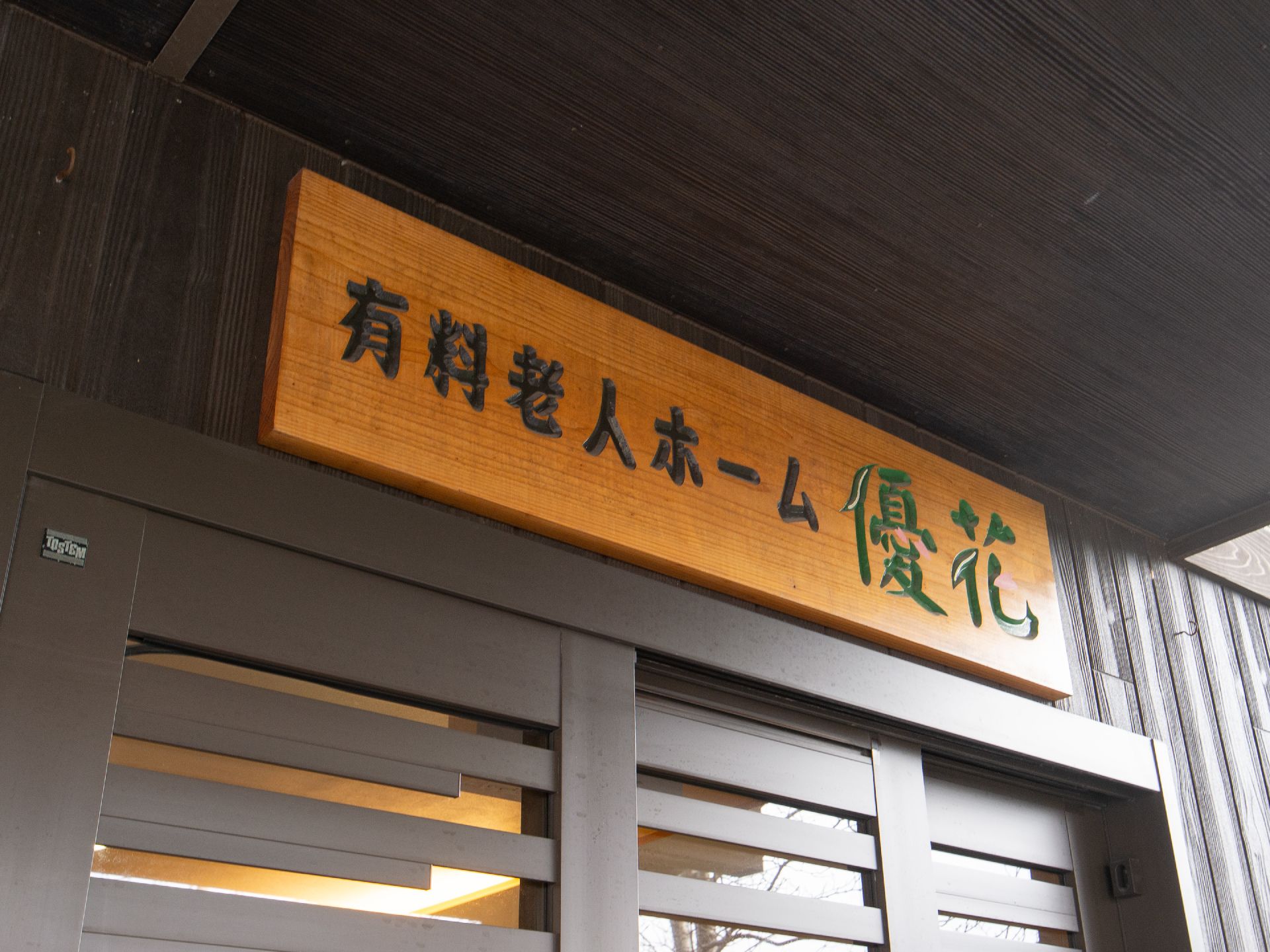 施設種別と施設名が彫刻された木製の看板が、玄関の上に掲げられている。洒落たデザインの文字が色つけされている。