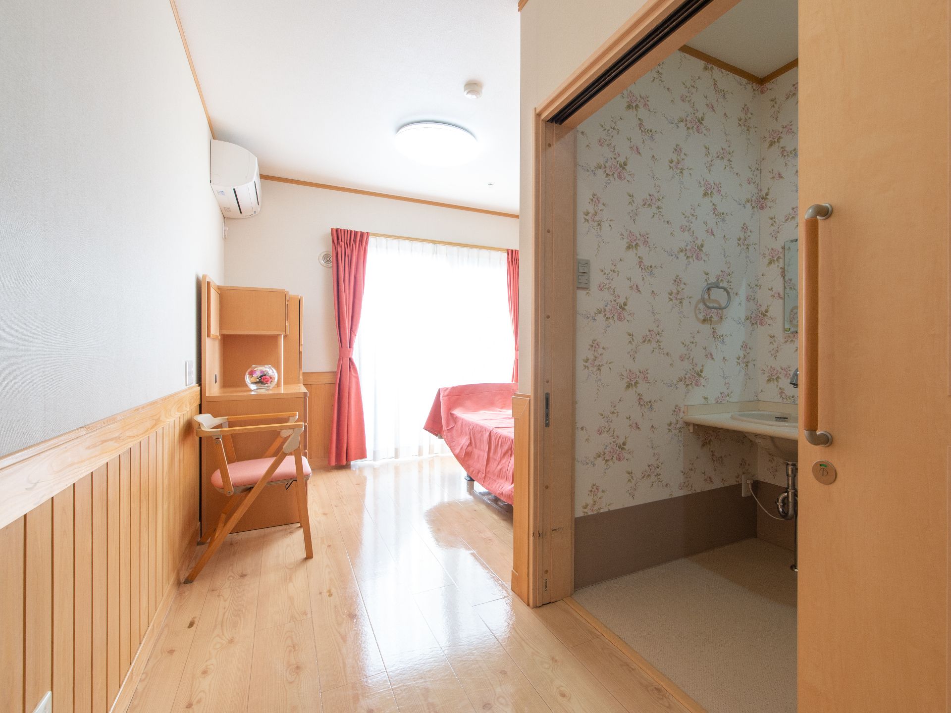 居室には大きな掃き出し窓がある。ピンク系のカーテンやクロスなどが使用されている。エアコンが完備されている。