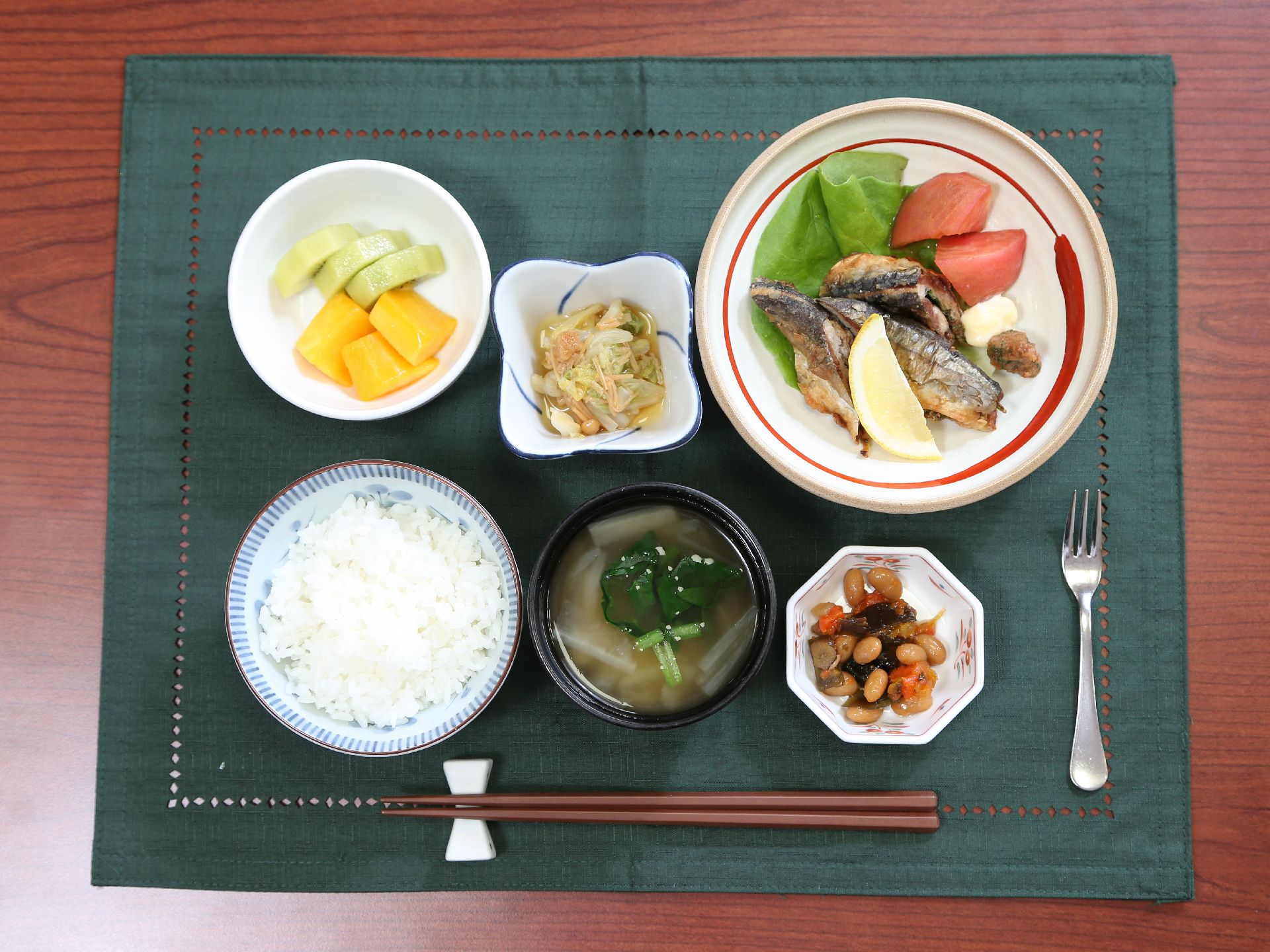 緑色のランチョンマットの上に食事が用意されている。焼き魚にトマト、煮物などの小鉢にご飯や味噌汁などが提供されている。