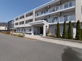 白色の３階建ての建物である。玄関前には車寄せがあり、屋根がある。建物の前にはさまざまな木が植えられている。