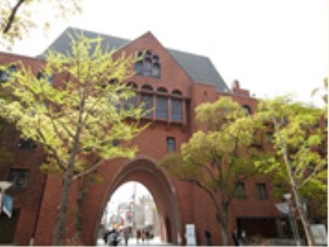 三角屋根の建物にアーチ形をした茶色の大きな門があり、多くの人々が行き交っている。門の前の大きな樹木が木陰を作ってくれている。