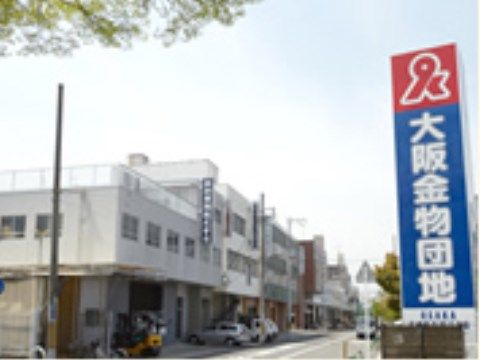 右の手前に団地のロゴと名称が書かれた縦長の看板が掲げてある。左手にはコンクリートの2階や３階建ての建物が連なっている。