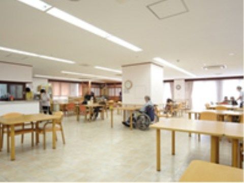 窓から光が差し込む、明るくて広い室内に家具がゆったり配置されている。テーブルの前には、車椅子や椅子に座る入居者様がおられる。