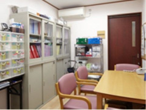 机の前には椅子が並んでいる。背後には書類棚が２台置かれ、他にも収納棚がある。整理が行き届き、すっきりした室内である。