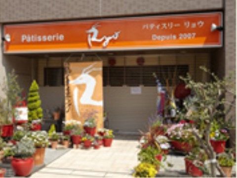 店の前はタイル張りで、多くの植物の鉢が並んでいる。花鉢のほか、ハーブやオリーブの木もある。オレンジ色のひさしが印象的。