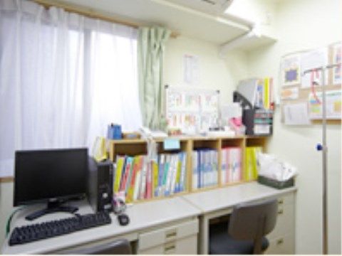 健康管理室には事務机が２つ並べられており、机の上には本やパソコンが置かれている。ガートルスタンドもある。