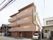 施設は４階建てで、茶色に近いベージュ色の外観である。敷地内には白い車が１台停められている。自動販売機もある。