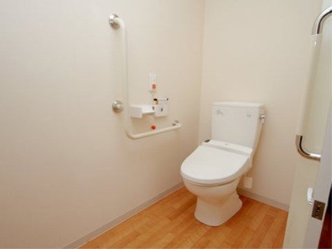 フローリングの床に白い壁紙の清潔感のあるトイレである。壁には、手すりや緊急時の呼び出しボタンが設置されている。