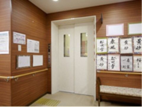 木目調の壁に設置されたエレベーターの白い扉が目を引くエレベーターホールである。壁にはさまざまな掲示物が貼られている。