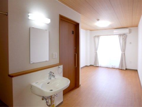 窓が大きく明るい居室。鏡のついたシンプルな洗面台とタオルかけが設置されている。居室内のトイレのドアは横開きで車椅子でも利用しやすい。