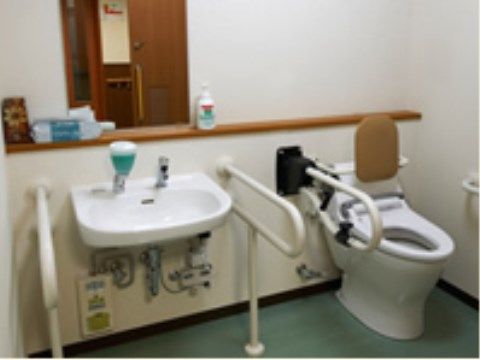 上に跳ね上げることができる可動式の手すりがついたトイレ。同じ室内には鏡のついたシンプルな手洗いシンクが設置されている。