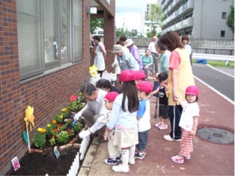 入居者様が幼稚園の子どもたちに花の植え方を教えている。花壇には黄色やピンクなどの花が植えられている。
