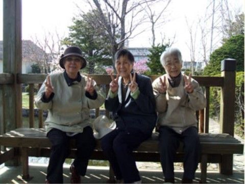 3人の入居者様が公園のベンチに座って笑顔でピースして写っている。周りには緑や花が多く植えられている。