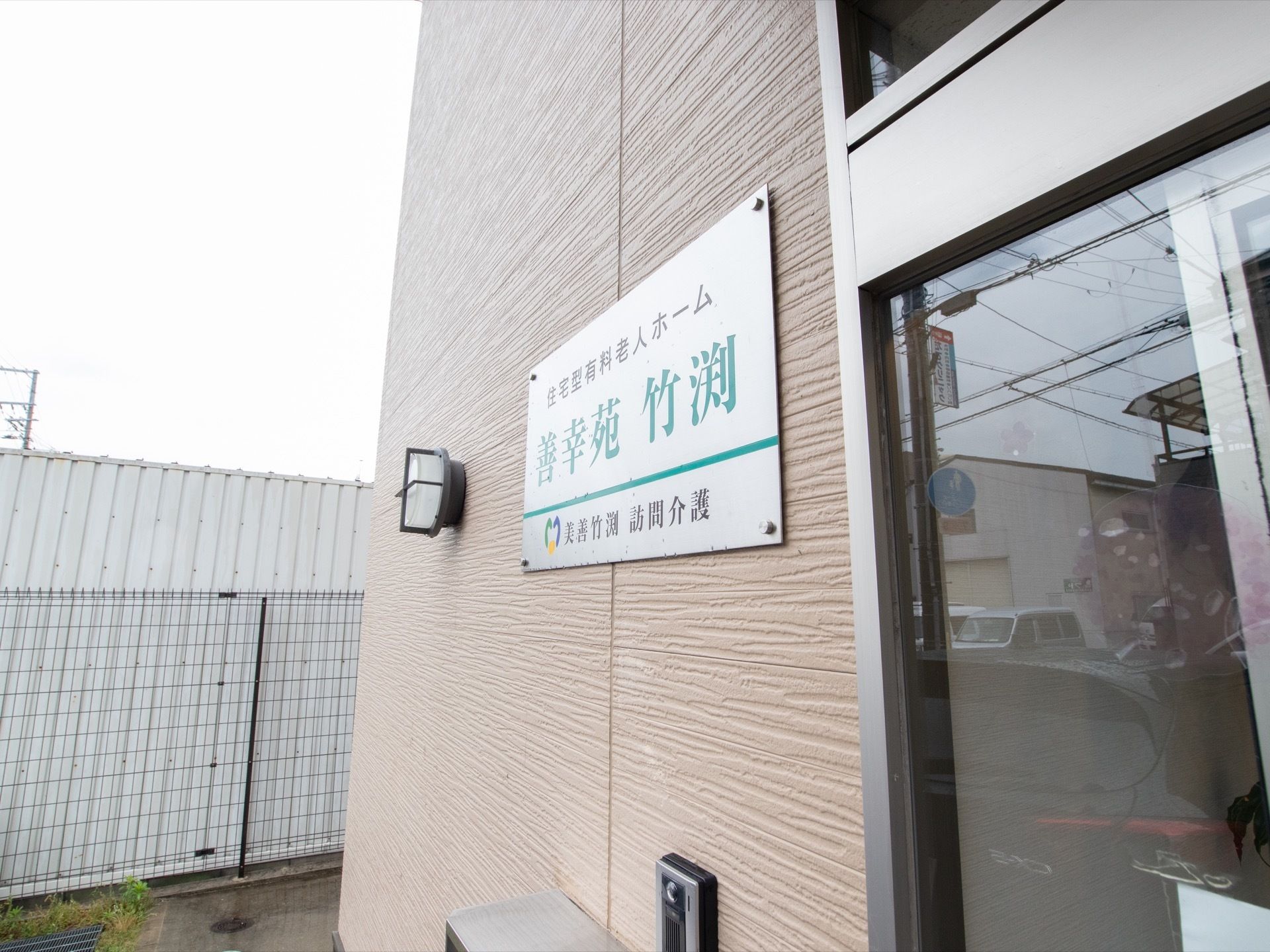 玄関の扉すぐ横の壁には施設の名称が書かれている金属製の看板がある。インターホンと外灯が設置されている。