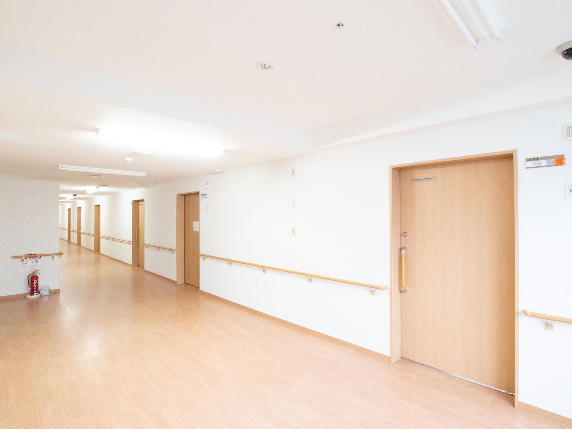 入居者様の個室が並ぶ廊下と広いホールがある。隅のわかりやすい場所に消火器が置かれており、壁には手すりが設置されている。