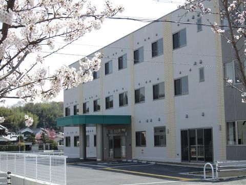 満開の桜の木の奥に建つ、白い壁に窓が並んだ３階建ての施設