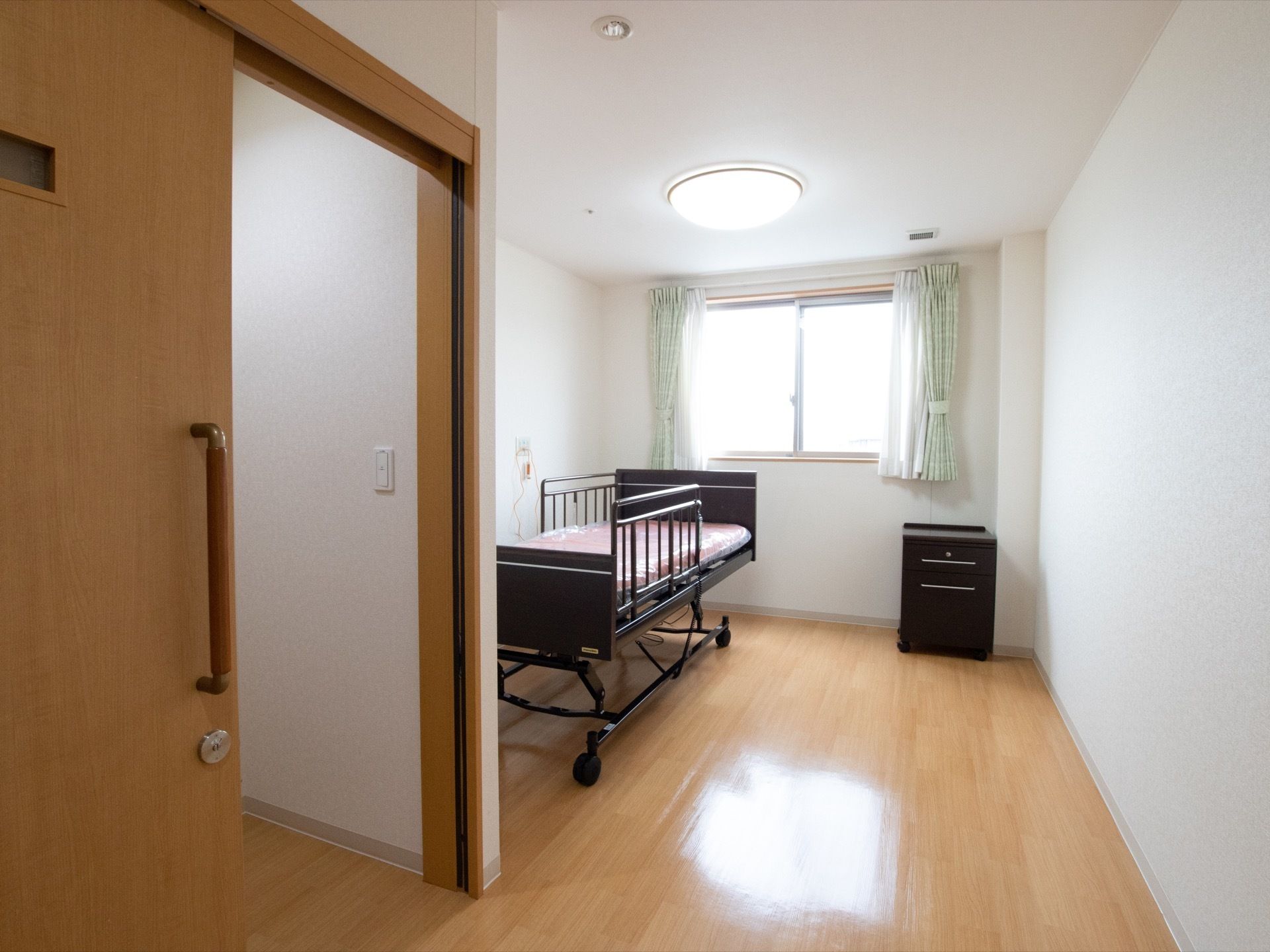 居室の窓には若草色のカーテンが下がっている。電動介護ベッドの色と同じこげ茶のチェストが置かれており、インテリアに統一感がでている。