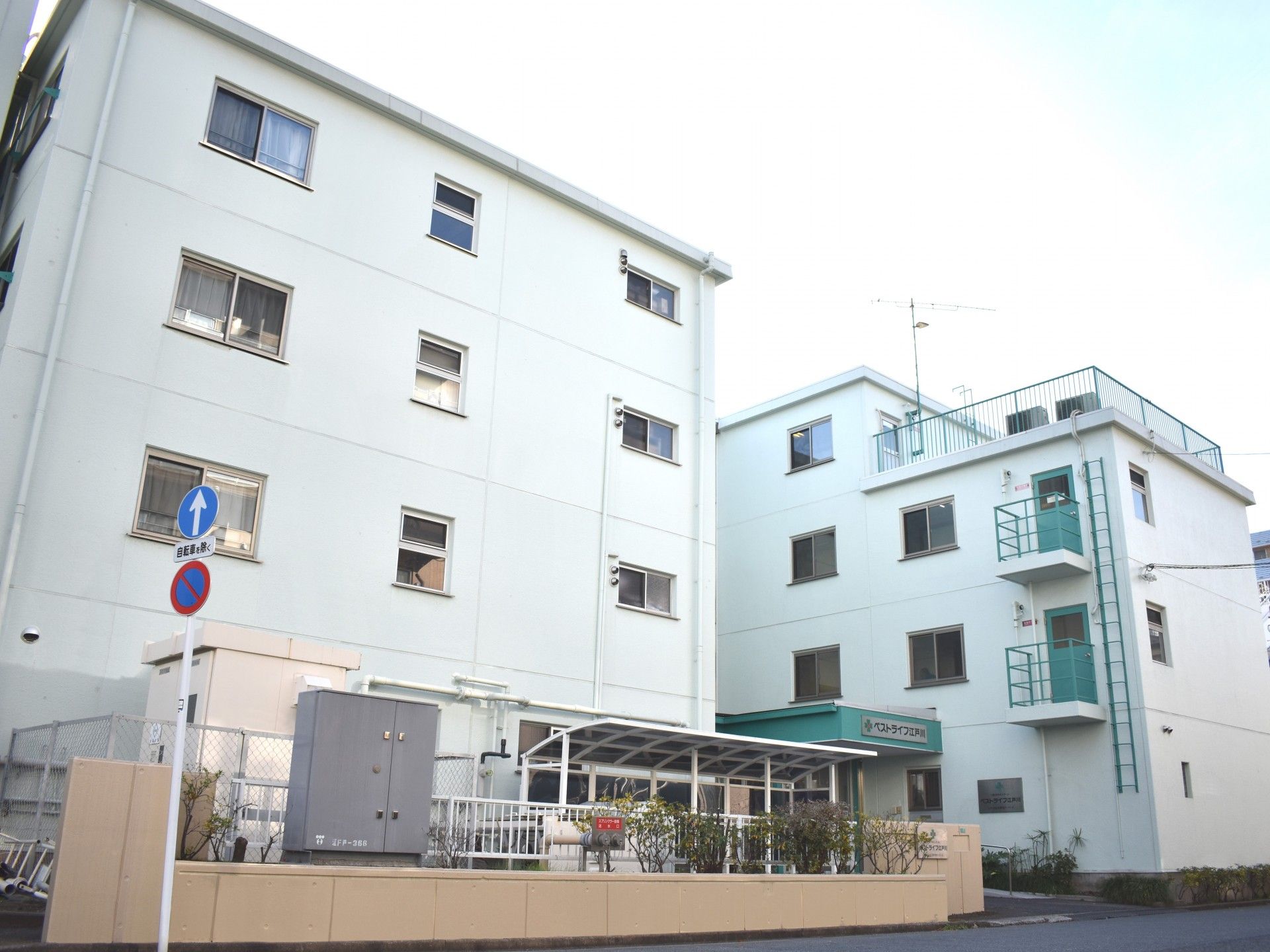 白い外壁が美しい、鉄筋4階建ての施設の外観。エントラスト横には駐輪場も設置されている。施設の周りには、緑が植えられている。