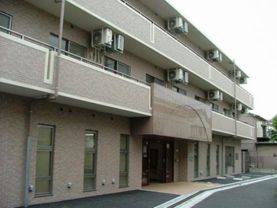 ベージュが基調の３階建ての建物で、１階には縦長の窓がいくつもある。入口の右側には数台分の駐車スペースがある。