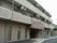 ベージュが基調の３階建ての建物で、１階には縦長の窓がいくつもある。入口の右側には数台分の駐車スペースがある。