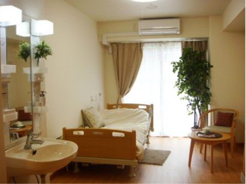 手前左側に洗面台がある。ベッドやテーブルなどがあり、ベッドの真ん中あたりに白いクッションが置かれている。